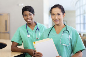 Smiling African American and caucasian nurse at hospital work station lit brightly with carts and wearing stethoscopes. Caucasian nurse in foreground.